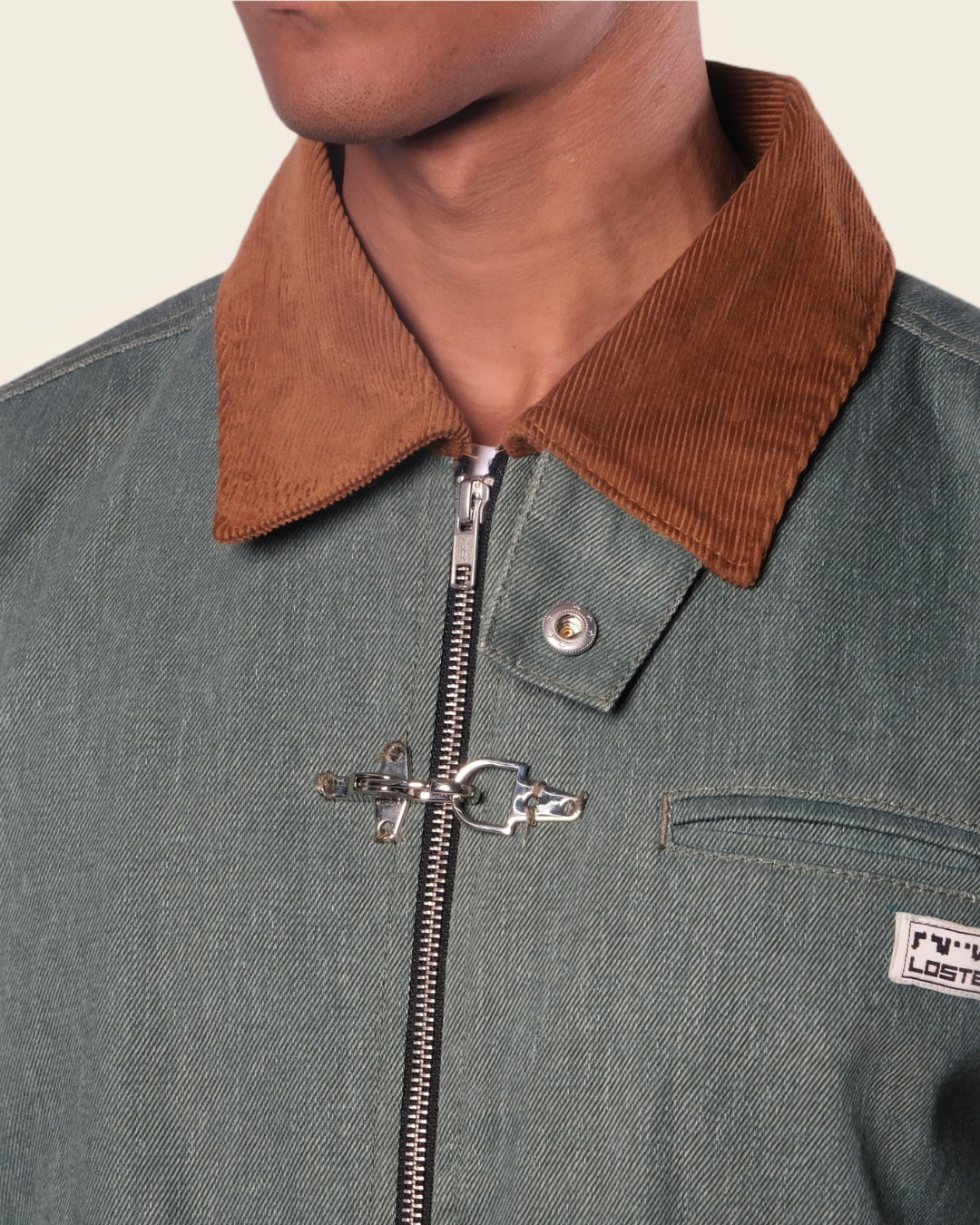 Close-up of a green jacket with a brown collar and zipper, featuring a brand logo on a beige background.