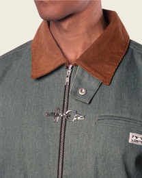 Close-up of a green jacket with a brown collar and zipper, featuring a brand logo on a beige background.
