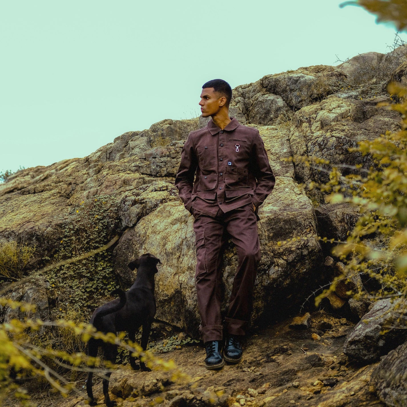 Man in a brown outfit standing on a rocky mountain with a dog beside him